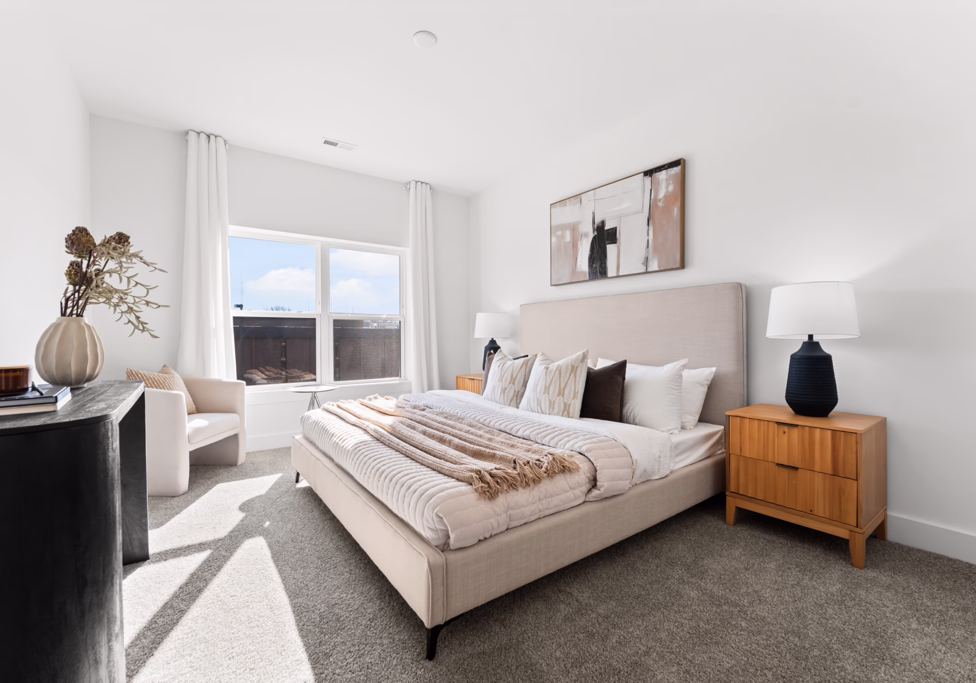 A carpeted bedroom with a made bed, wooden nightstand and afternoon light from a tall window.
