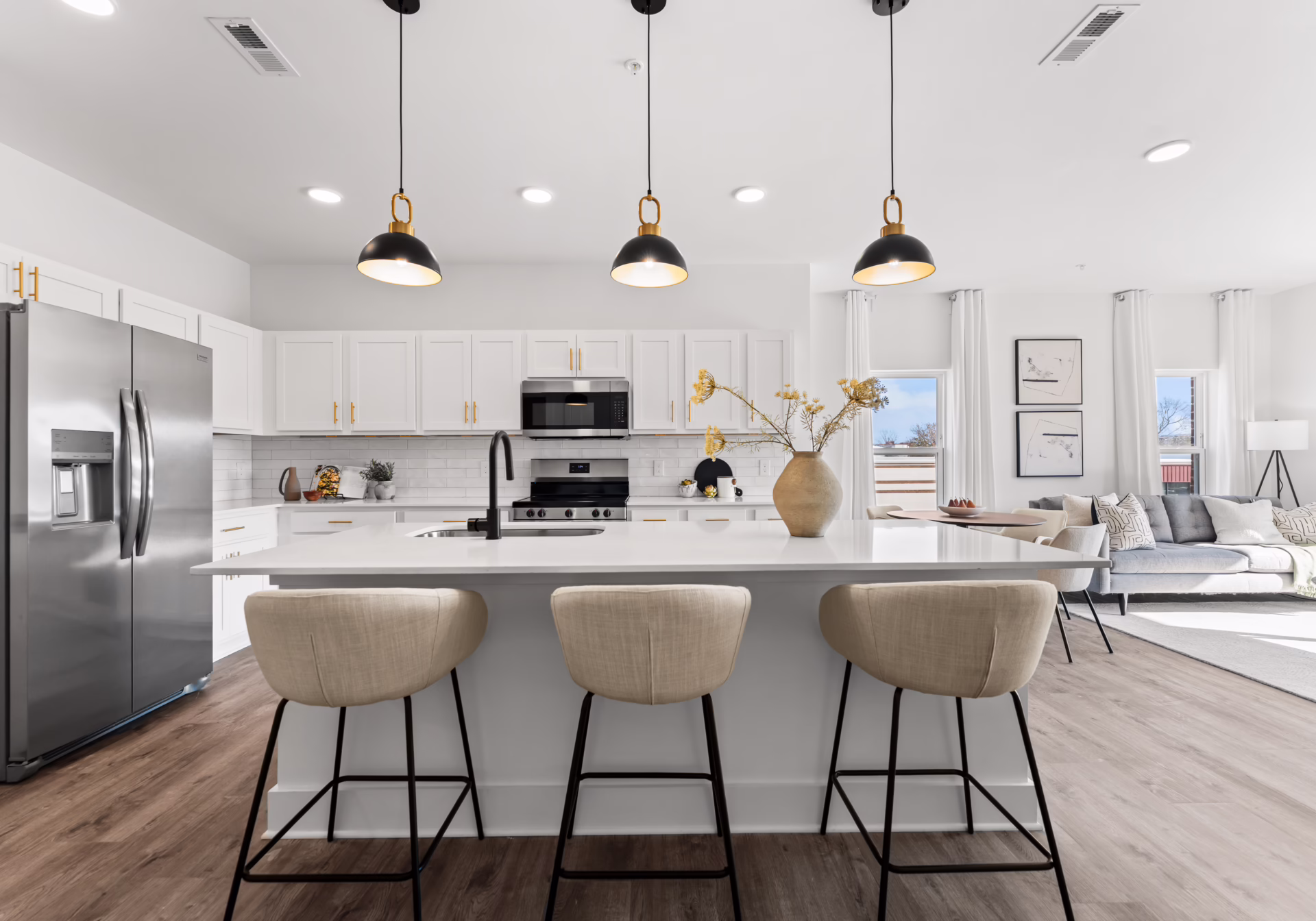 Kitchen with a white quartz island and three barstools, white shaker cabinets with brass pulls, stainless appliances and matte-black pendant lights.